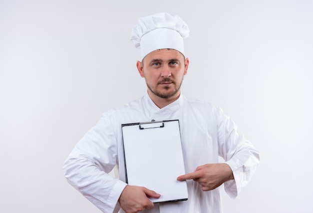 Zelfverzekerde jonge knappe kok in chef-kok uniform houden en wijzend op klembord geïsoleerd op een witte muur