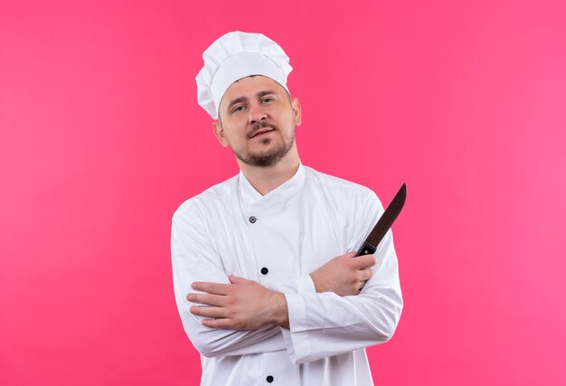 Zelfverzekerde jonge knappe kok in chef-kok uniform staande met gesloten houding en met mes geïsoleerd op roze muur