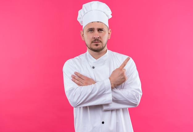 Zelfverzekerde jonge knappe kok in uniform van de chef-kok die de hand op de arm legt en naar de zijkant wijst, geïsoleerd op een roze muur
