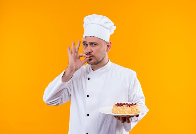 Zelfverzekerde jonge knappe kok in uniform van de chef-kok die een bord cake vasthoudt en een smakelijk gebaar doet geïsoleerd op een oranje muur