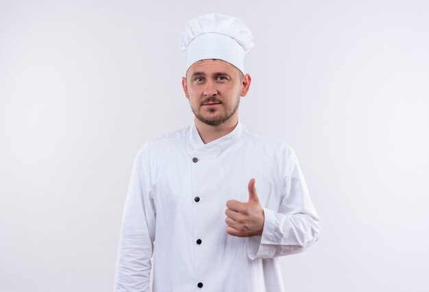 Zelfverzekerde jonge knappe kok in uniform van de chef-kok met duim omhoog geïsoleerd op een witte muur