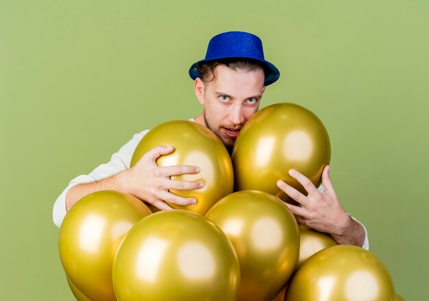 Zelfverzekerde jonge knappe Slavische feestmens met feestmuts achter ballonnen grijpen ze kijken naar camera geïsoleerd op olijfgroene achtergrond