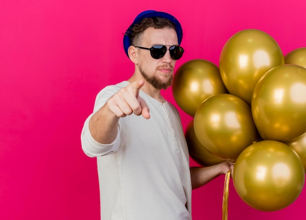 Zelfverzekerde jonge knappe Slavische feestmens met feestmuts en zonnebril met ballonnen kijken en wijzend naar voorzijde geïsoleerd op roze muur met kopie ruimte