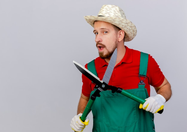 Zelfverzekerde jonge knappe Slavische tuinman in uniform en hoed met snoeischaar geïsoleerd op een witte muur met kopie ruimte