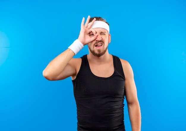 Zelfverzekerde jonge knappe sportieve man met hoofdband en polsbandjes die een gebaar maken dat op een blauwe muur wordt geïsoleerd