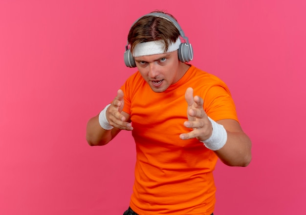 Zelfverzekerde jonge knappe sportieve man met hoofdband en polsbandjes en koptelefoon handen geïsoleerd op roze met kopie ruimte strekken