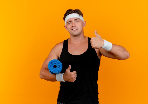 Zelfverzekerde jonge knappe sportieve man met hoofdband en polsbandjes houden handdoek en duimen opdagen geïsoleerd op oranje met kopie ruimte