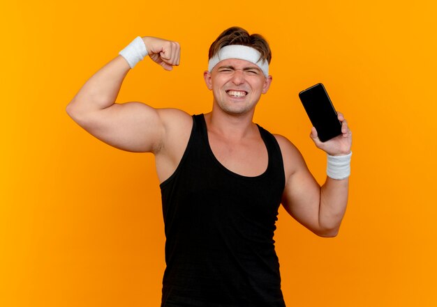 Zelfverzekerde jonge knappe sportieve man met hoofdband en polsbandjes met mobiele telefoon en gebaren sterk geïsoleerd op oranje
