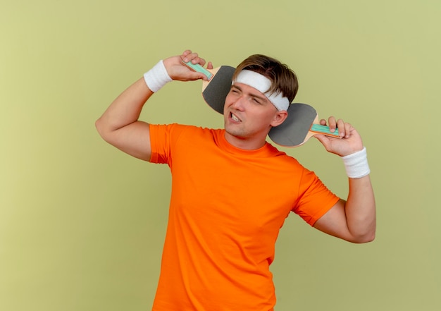 Zelfverzekerde jonge knappe sportieve man met hoofdband en polsbandjes met pingpongrackets achter hoofd kijken naar kant geïsoleerd op olijfgroen