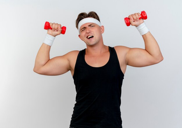 Gratis foto zelfverzekerde jonge knappe sportieve man met hoofdband en polsbandjes verhogen halters geïsoleerd op wit