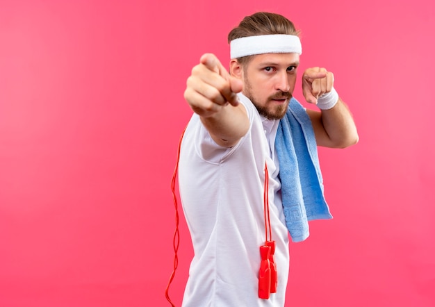 Zelfverzekerde jonge knappe sportieve man met hoofdband en polsbandjes wijzend met springtouw en handdoek op schouders geïsoleerd op roze muur met kopieerruimte