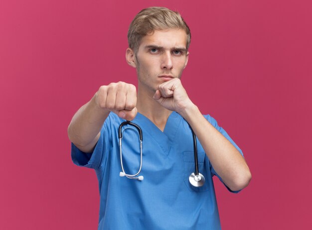 Zelfverzekerde jonge mannelijke arts die doktersuniform draagt met een stethoscoop die staat in de strijd, geïsoleerd op roze muur