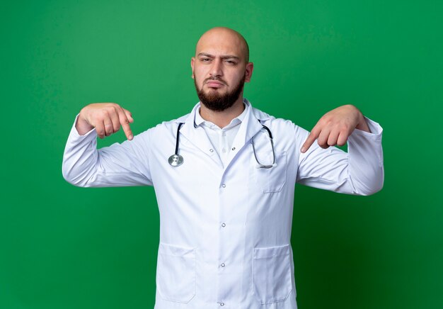 Zelfverzekerde jonge mannelijke arts die medische mantel en stethoscoop draagt, wijst naar zichzelf geïsoleerd op groene achtergrond