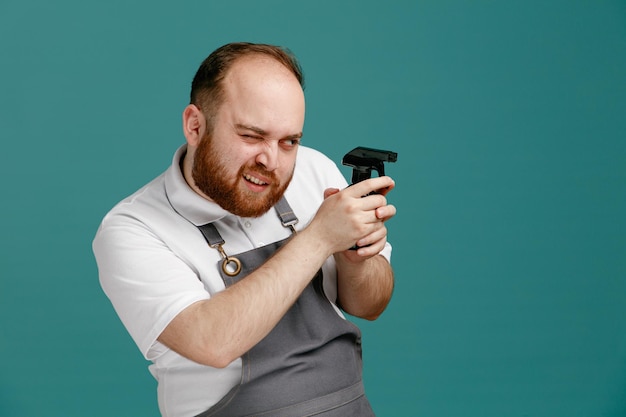 Zelfverzekerde jonge mannelijke kapper met een wit overhemd en een kappersschort die naar de zijkant kijkt met één oog gesloten en een spuitfles vasthoudt met beide handen geïsoleerd op een blauwe achtergrond met kopieerruimte