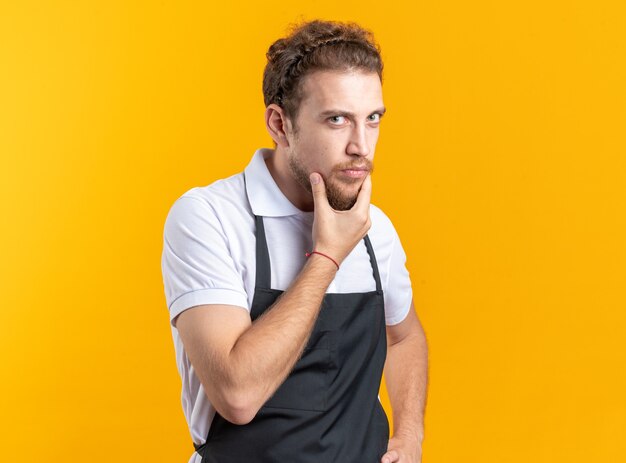 Zelfverzekerde jonge mannelijke kapper met uniforme greep kin geïsoleerd op gele muur