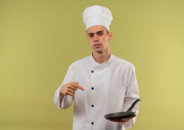 Zelfverzekerde jonge mannelijke kok die chef-kok uniform draagt, wijst vinger naar koekenpan in zijn hand op geïsoleerde groene muur met kopie ruimte