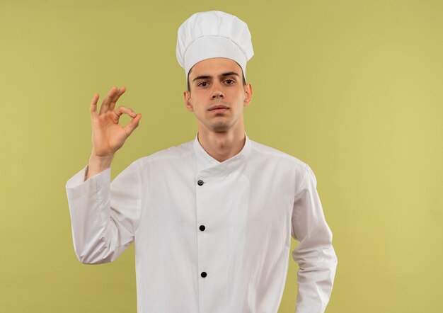 Zelfverzekerde jonge mannelijke kok die eenvormige chef-kok draagt die okgebaar op geïsoleerde groene muur met exemplaarruimte toont