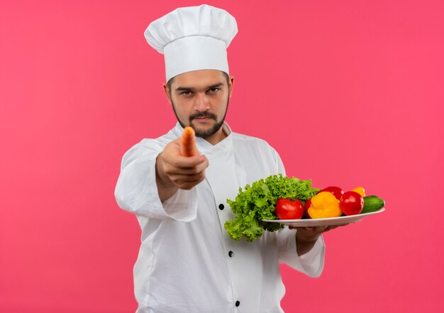 Zelfverzekerde jonge mannelijke kok in chef-kok uniform met bord groenten en wijzend met wortel geïsoleerd op roze muur met kopieerruimte copy