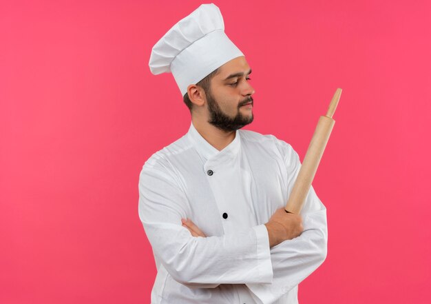 Zelfverzekerde jonge mannelijke kok in chef-kok uniform staande met gesloten houding en deegroller vasthoudend kijkend naar kant geïsoleerd op roze muur met kopieerruimte
