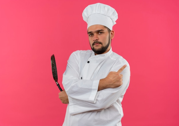 Zelfverzekerde jonge mannelijke kok in uniform van de chef-kok die een koekenpan vasthoudt en naar de zijkant wijst, geïsoleerd op een roze muur met kopieerruimte