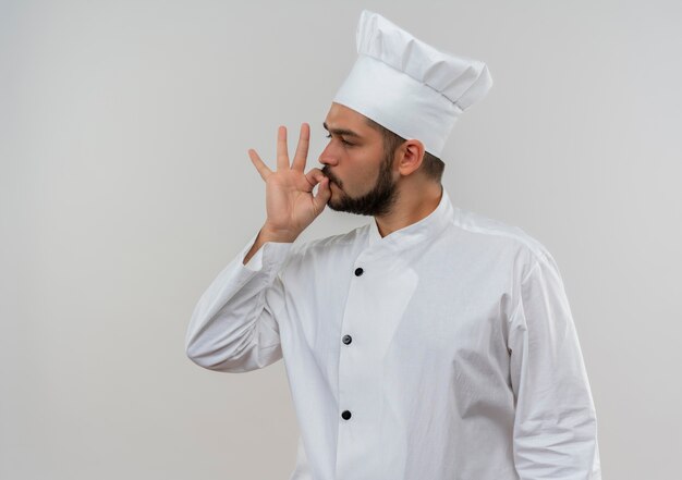 Zelfverzekerde jonge mannelijke kok in uniform van de chef-kok die naar de zijkant kijkt en een smakelijk gebaar doet dat op een witte muur met kopieerruimte wordt geïsoleerd