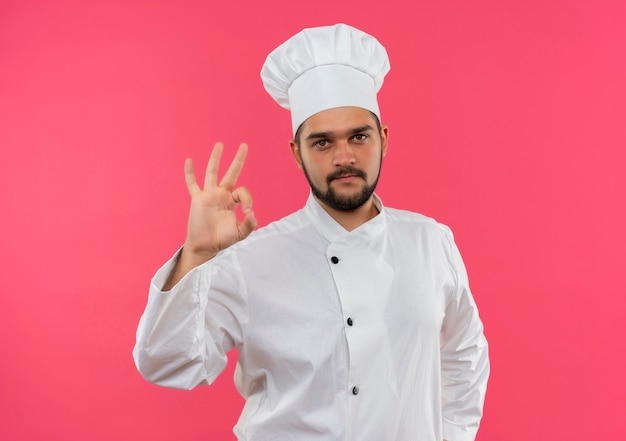 Zelfverzekerde jonge mannelijke kok in uniform van de chef-kok doet ok teken geïsoleerd op roze muur