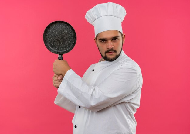 Zelfverzekerde jonge mannelijke kok in uniform van de chef-kok met koekenpan geïsoleerd op roze muur