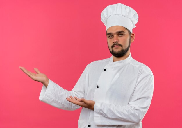 Zelfverzekerde jonge mannelijke kok in uniform van de chef-kok wijzend met de handen aan de zijkant geïsoleerd op roze muur