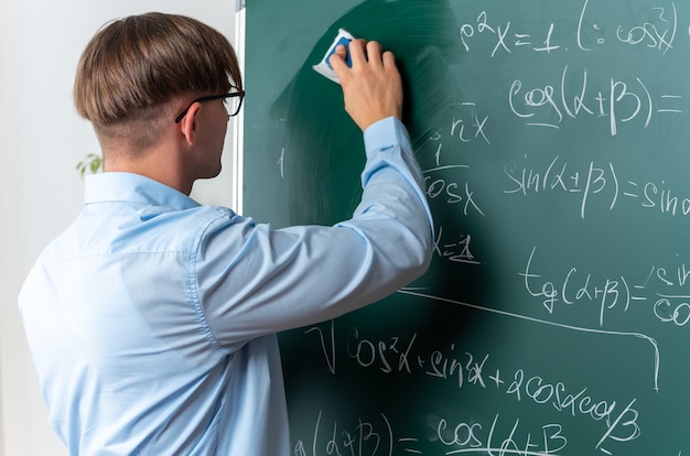 Zelfverzekerde jonge mannelijke leraar met een bril die bij het bord staat en wiskundige formules in de klas afveegt