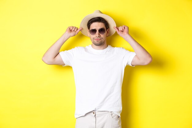Zelfverzekerde jonge mannelijke toerist klaar voor vakantie, met strohoed en zonnebril, staande tegen een gele achtergrond.