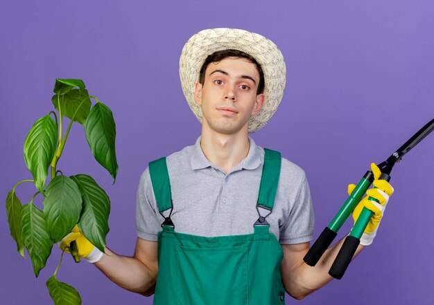 Zelfverzekerde jonge mannelijke tuinman tuinieren hoed en handschoenen houdt plant en tondeuse