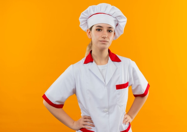 Zelfverzekerde jonge mooie kok in chef-kok uniform handen op taille geïsoleerd op oranje muur zetten