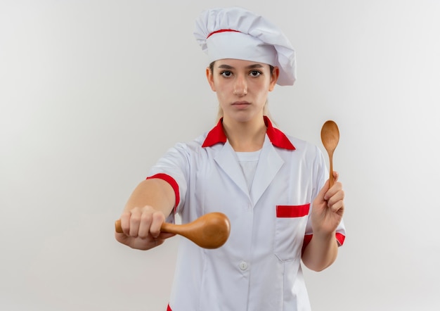 Zelfverzekerde jonge mooie kok in uniform van de chef-kok die lepels vasthoudt en uitrekt die op een witte muur zijn geïsoleerd