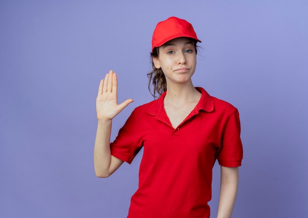 zelfverzekerde jonge mooie leveringsvrouw die rode uniform en pet draagt die hallo gebaar doet