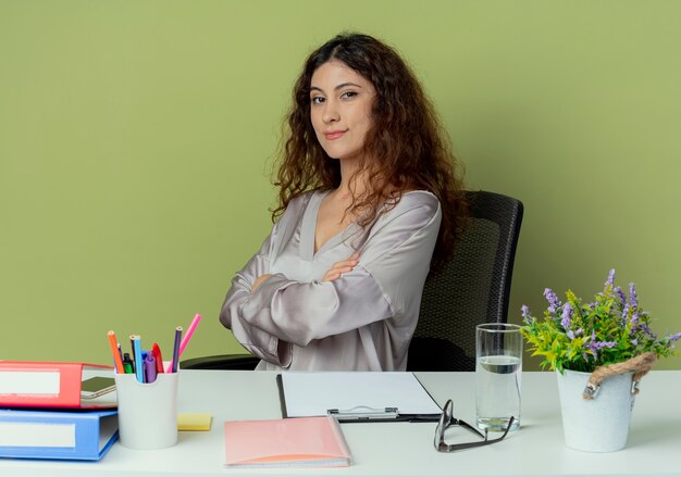 Zelfverzekerde jonge mooie vrouwelijke beambte zittend aan een bureau met kantoorhulpmiddelen die handen kruisen die op olijfachtergrond worden geïsoleerd