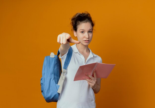 Zelfverzekerde jonge mooie vrouwelijke student die achterzak draagt die open notitieblok houdt en op camera richt die op oranje achtergrond met exemplaarruimte wordt geïsoleerd