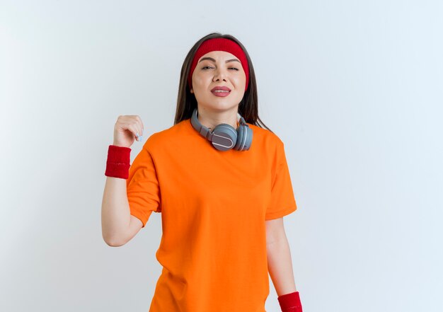 Zelfverzekerde jonge sportieve vrouw met hoofdband en polsbandjes en koptelefoon op nek houden hand in lucht geïsoleerd op een witte muur met kopie ruimte