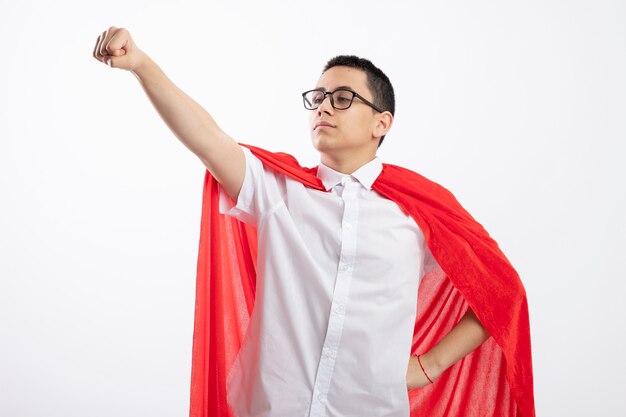 Zelfverzekerde jonge superheld jongen in rode cape bril houden hand op taille kijken kant strekken vuist geïsoleerd op een witte achtergrond