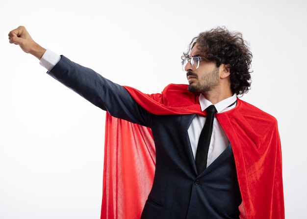Gratis foto zelfverzekerde jonge superheld man in optische bril dragen pak met rode mantel werpt vuist omhoog en kijkt naar kant geïsoleerd op witte muur