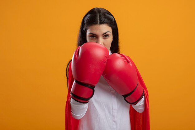 Zelfverzekerde jonge supervrouw die dooshandschoenen draagt die voorzijde bekijken die handen samen houden die op oranje muur worden geïsoleerd