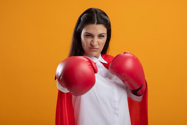 Zelfverzekerde jonge supervrouw die dooshandschoenen draagt die voorzijde bekijkt die boksgebaar doet dat op oranje muur wordt geïsoleerd