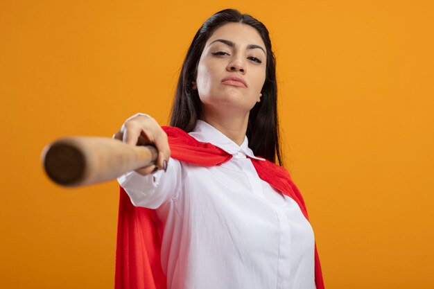 Zelfverzekerde jonge superwoman die honkbalknuppel uitrekt naar voorzijde die voorzijde bekijkt die op oranje muur wordt geïsoleerd