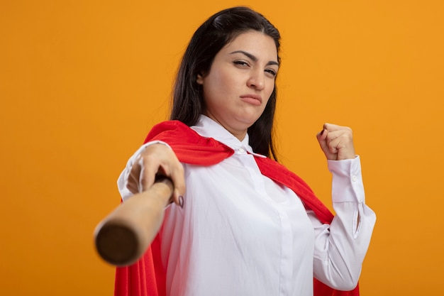 Zelfverzekerde jonge superwoman honkbalknuppel uitrekken naar voren kijken naar de voorkant gebalde vuist geïsoleerd op een oranje muur
