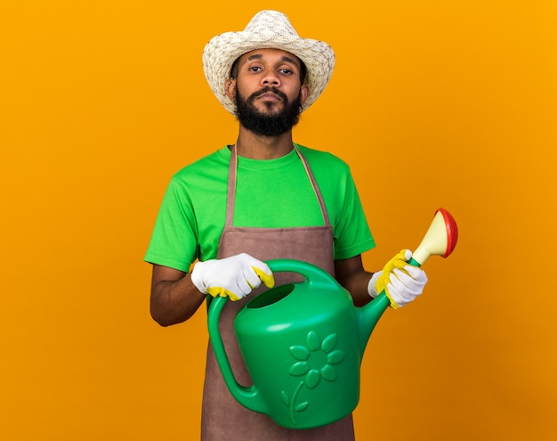 Zelfverzekerde jonge tuinman Afro-Amerikaanse man met een tuinhoed en handschoenen met een gieter geïsoleerd op een oranje muur