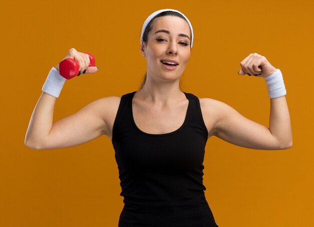 Zelfverzekerde jonge vrij sportieve vrouw met hoofdband en polsbandjes met halter die naar de voorkant kijkt en een sterk gebaar doet dat op een oranje muur wordt geïsoleerd