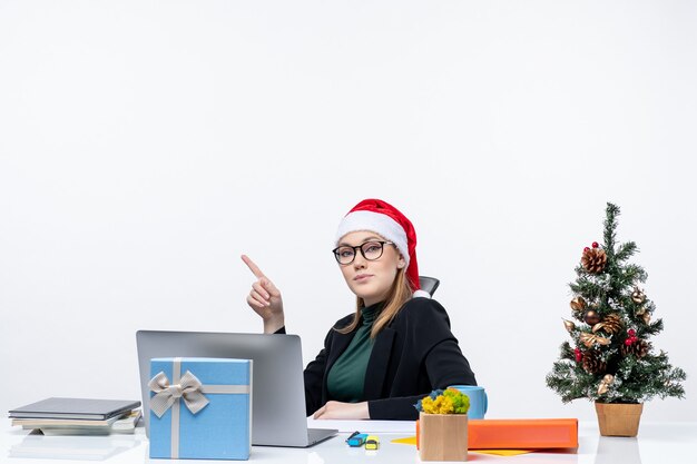 Zelfverzekerde jonge vrouw met kerstman hoed en bril zittend aan een tafel met een kerstboom en een cadeau erop op witte achtergrond