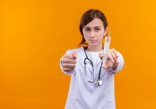 Zelfverzekerde jonge vrouwelijke arts die medische mantel en stethoscoop draagt die spuit en vuist uitrekt op geïsoleerde oranje ruimte met exemplaarruimte