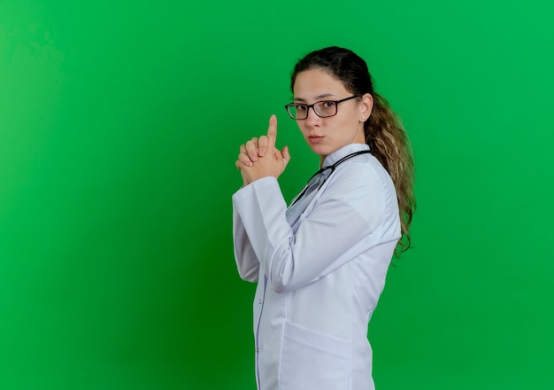 Zelfverzekerde jonge vrouwelijke arts die medische mantel en stethoscoop en bril draagt die zich in profielweergave bevindt die pistoolgebaar doet geïsoleerd op groene muur met exemplaarruimte