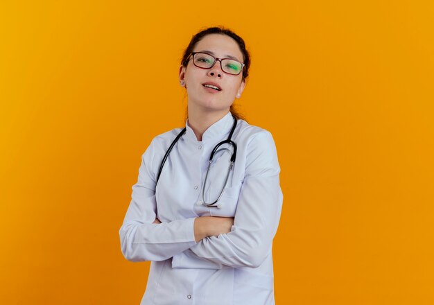 Zelfverzekerde jonge vrouwelijke arts die medische mantel en stethoscoop met bril draagt die geïsoleerde handen kruist