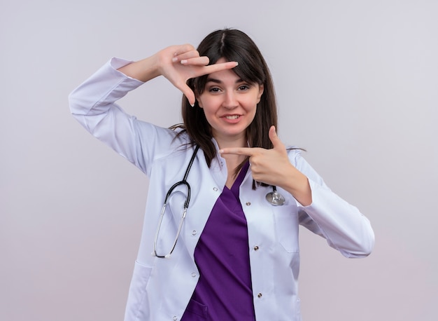 Zelfverzekerde jonge vrouwelijke arts in medische mantel met het frame van stethoscoopgebaren op geïsoleerde witte achtergrond